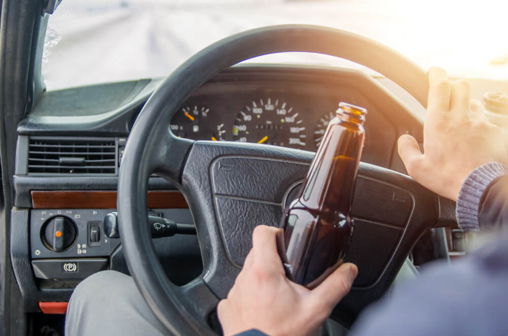 A drunken man driving a car with a bottle of alcohol in his hand.A man holds a driving wheel and a bottle of beer.