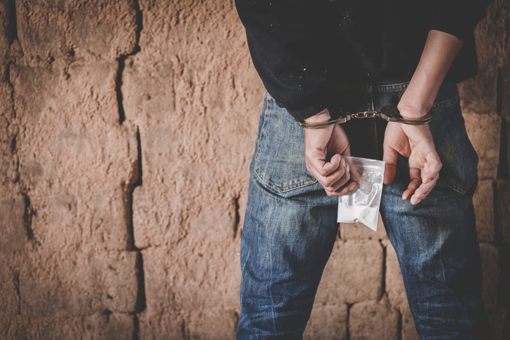 man standing facing brick wall with hands cuffed behind his back holding bag of drugs